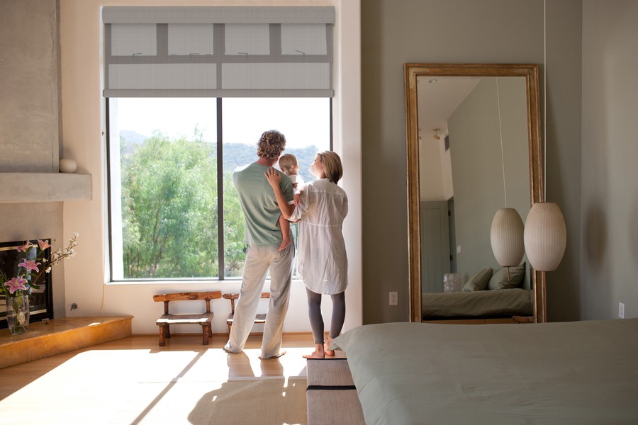 A family of three stands in front of a floor-to-ceiling window with partially lowered motorized shades. Green trees are in the distance.
