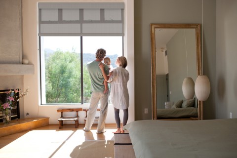 A family of three stands in front of a floor-to-ceiling window with partially lowered motorized shades. Green trees are in the distance.