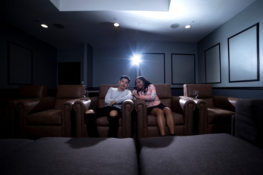 Two people sitting in plush home theater seats, eating popcorn. The glow of an unseen movie screen illuminates their faces.
